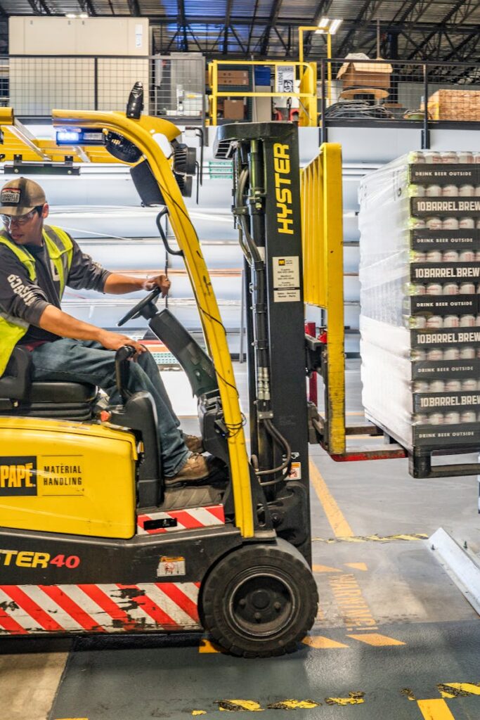 A warehouse worker maneuvers a forklift to transport crates for brewing company storage.